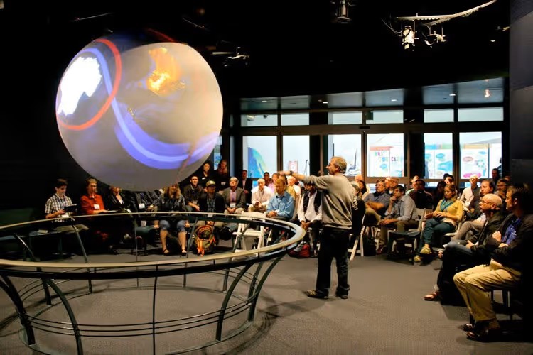 Person presenting an SOS exhibit with the SOS globe hanging from the ceiling and a rail around it. Lots of people in the back learning about SOS