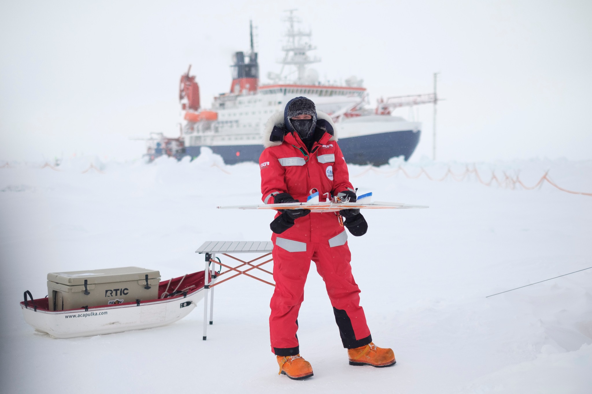 Person standing on an arctic ice field in a red suit with a boat behind them 
