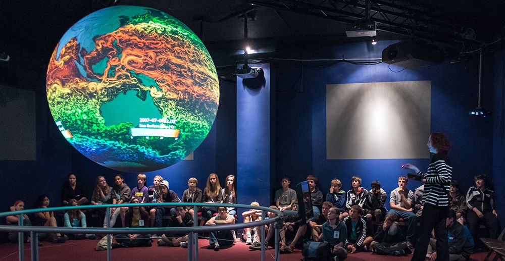 Beth Russel giving an SOS presentation to a large crowd of children. The SOS globe is hanging from the ceiling showing sea surface temperatures-green, blue, orange and yellow are shown on the globe