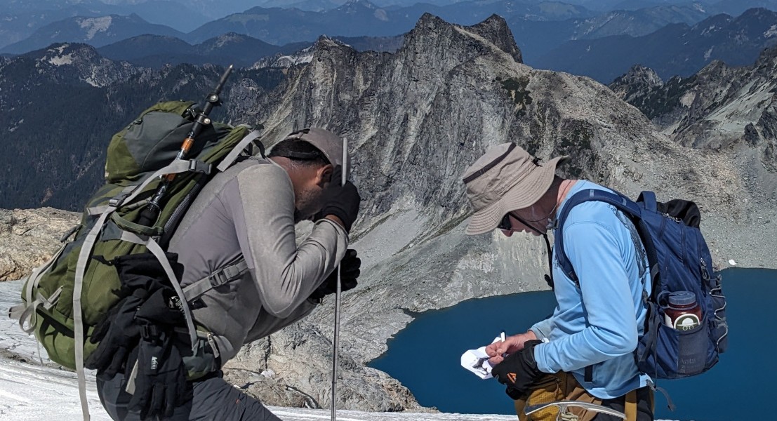 Two people conducting fieldwork on a mountain side with a blue lake below