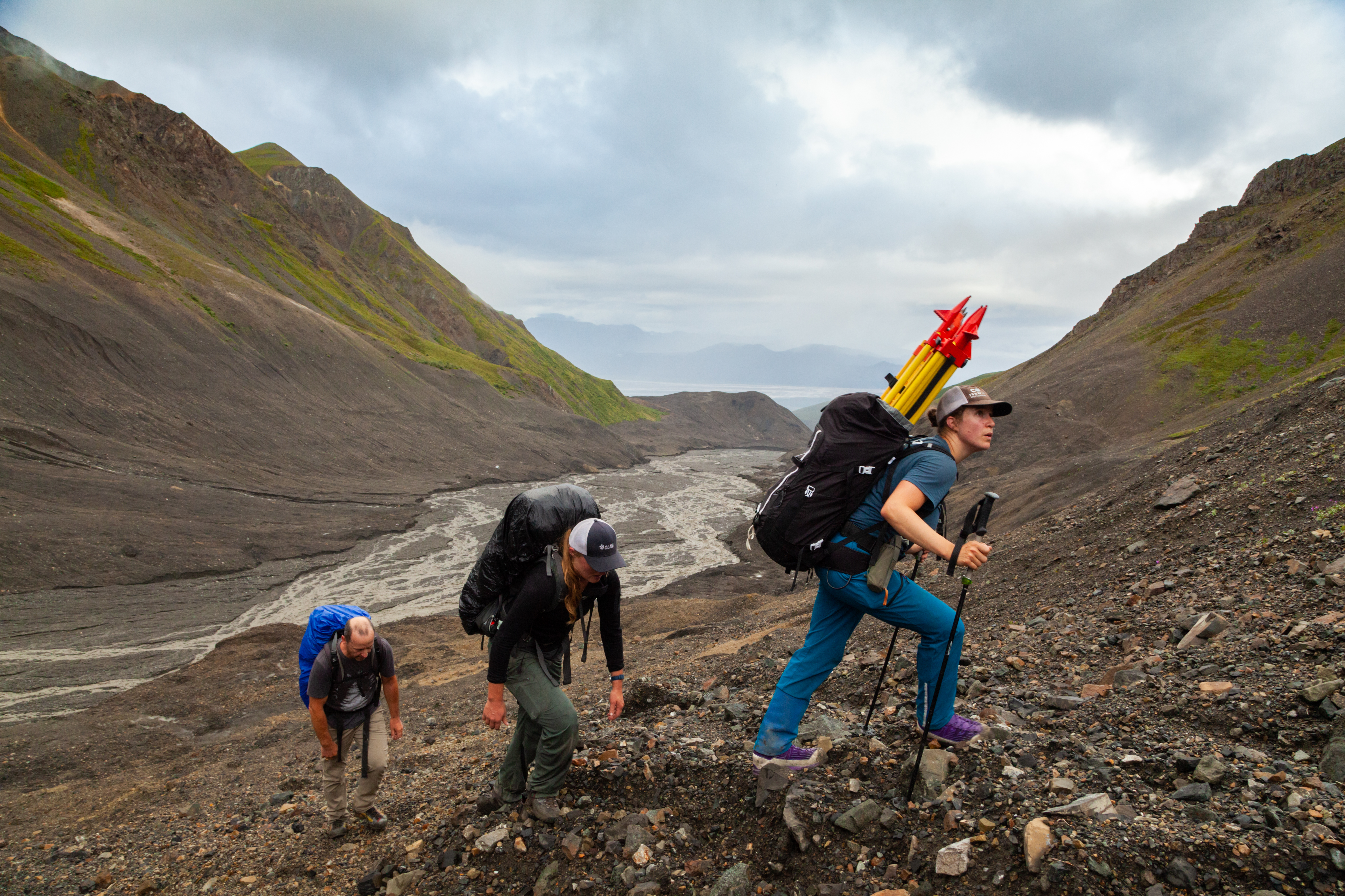Three hikers with large backpacks ascend a rocky, barren slope in a rugged mountain valley. The lead hiker carries bright yellow and red equipment, possibly for surveying or research. The terrain is steep and scattered with loose stones, with a glacial stream winding through the valley below. The sky is overcast, adding a dramatic feel to the remote and harsh landscape.