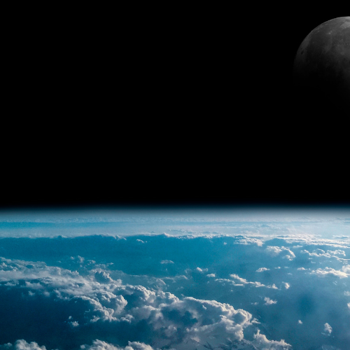  A view from space showing the Earth's curved horizon covered in a layer of clouds, with the dark expanse of space above. The Moon is partially visible in the upper right corner against the black sky.