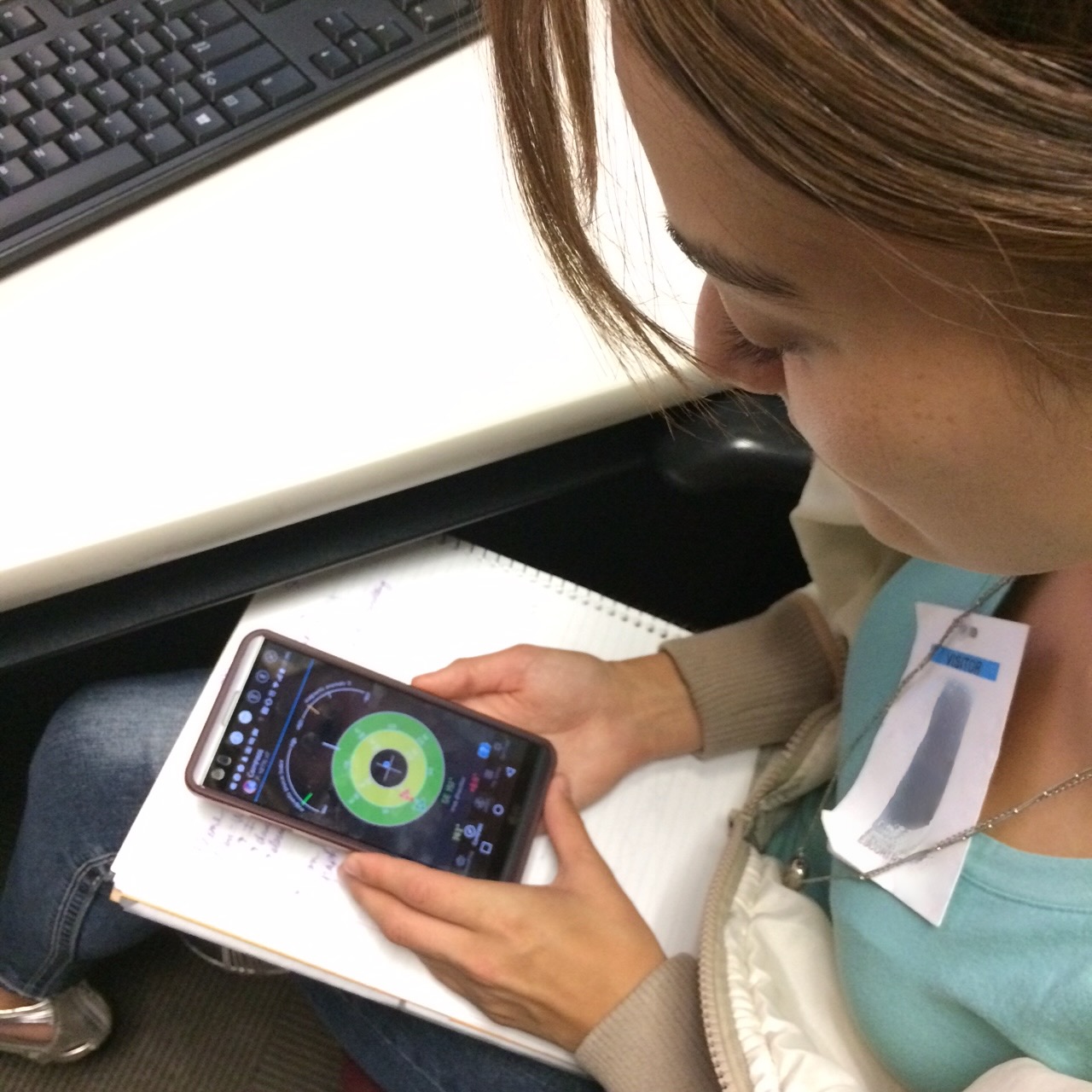 A person sitting at a desk looks at a smartphone displaying a colorful circular radar-like interface, possibly related to GPS or satellite tracking. The individual holds the phone over a notepad and wears a visitor badge with a blurred name tag. A computer keyboard is visible on the desk nearby.