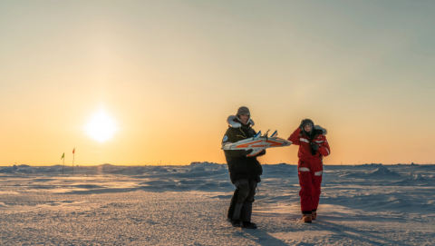 Two people wearing heavy winter gear stand on a snowy landscape at sunset. One person, dressed in black, is holding a guitar, while the other, dressed in red, is listening and smiling. The sky is clear with the sun low on the horizon, casting a warm golden glow across the icy terrain