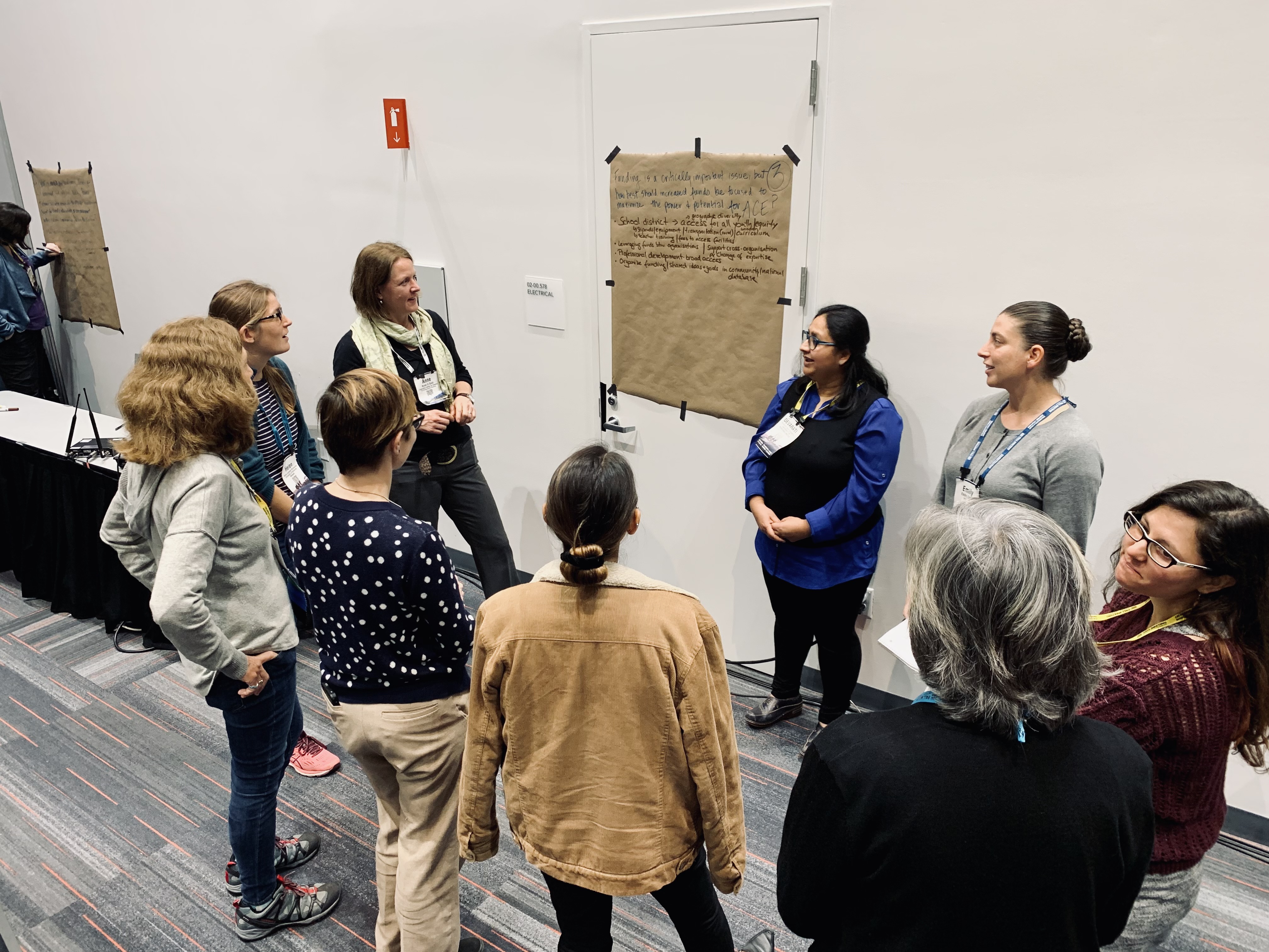 A group of women are gathered in a conference or workshop setting, standing in a circle around a large piece of brown paper taped to a white wall. The paper contains handwritten notes, and two women appear to be leading the discussion. The room has gray carpeting with red and blue lines, and there are more posters on the walls. The participants appear engaged and are wearing name tags and casual attire