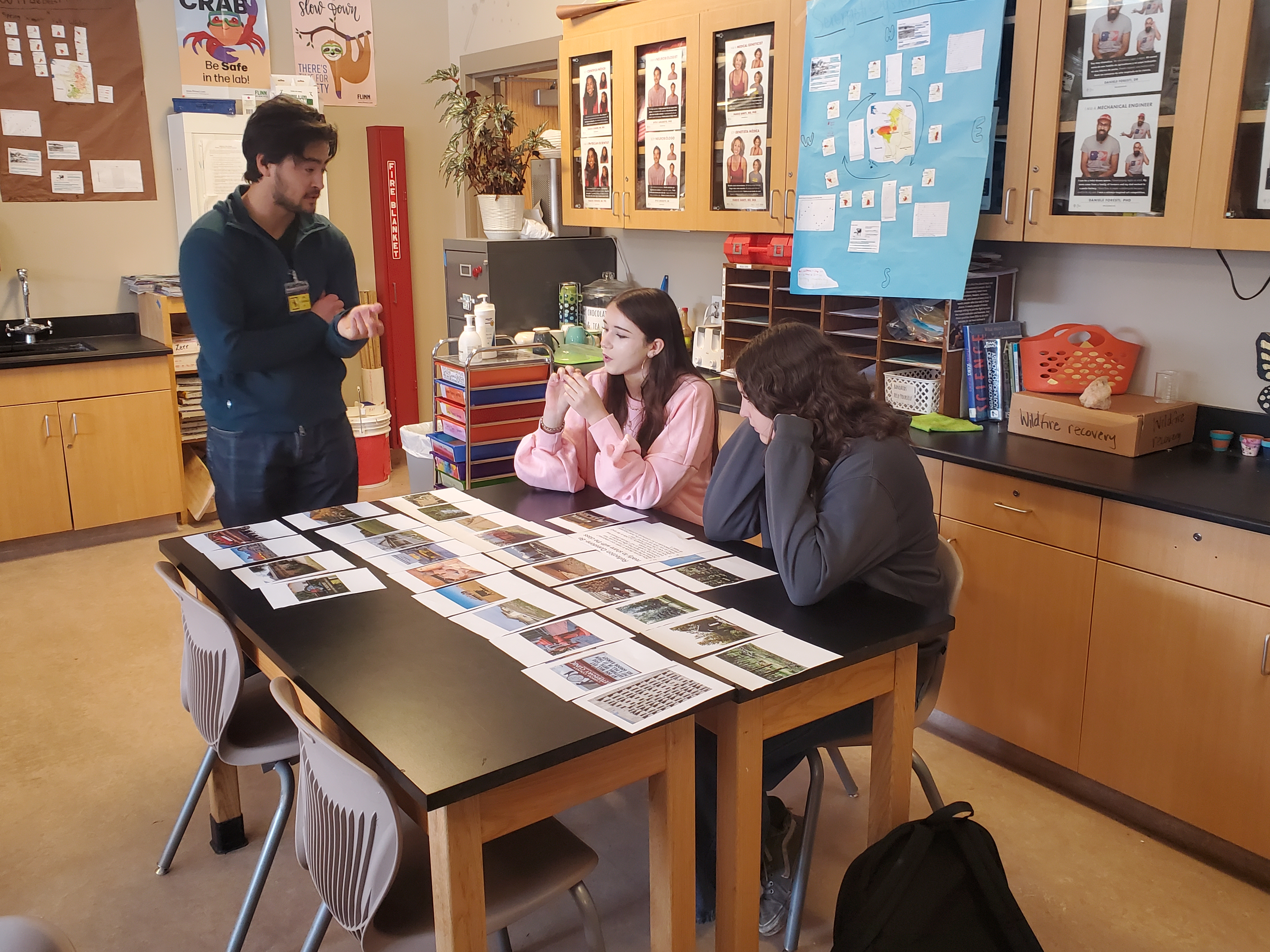 Two students sit at a classroom table covered with photo cards and printed materials while a teacher stands nearby explaining an activity. The students appear engaged, one leaning forward with hands clasped, the other resting her head on her hand. Behind them, cabinets display posters, and a large blue poster board with papers and diagrams hangs on the wall