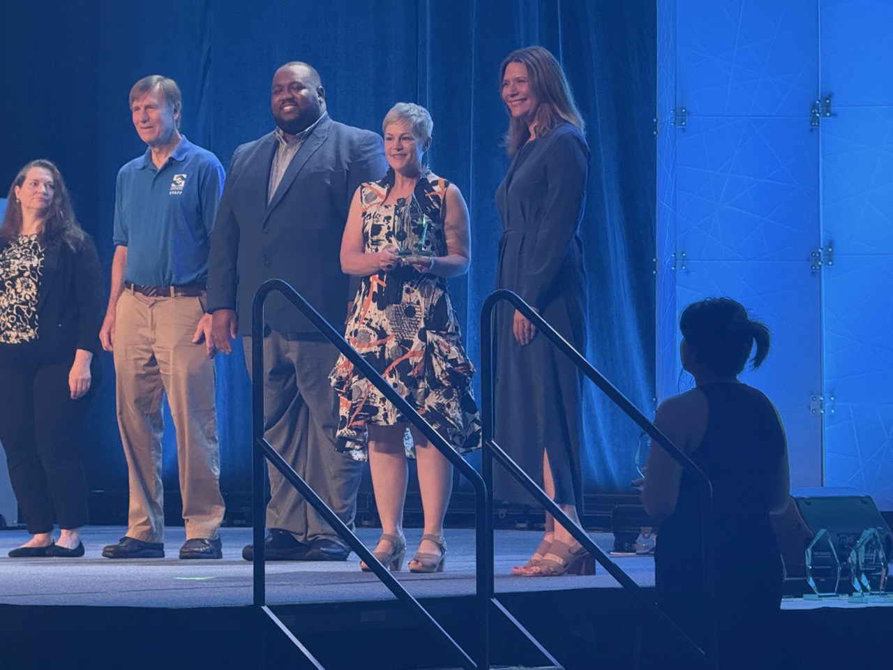 Carrie McDougall stands on stage with peers and award presenters at the ASTC Award Ceremony as she is presented with her trophy celebrating her achievement.