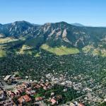 Aerial view of Boulder, Colorado, showing the city with its grid-like streets surrounded by lush greenery, with the Flatirons rock formations and mountains in the background under a clear blue sky