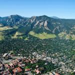 Aerial view of Boulder, Colorado, showcasing a sprawling cityscape nestled against the dramatic backdrop of the Flatirons—steep, slanted sandstone rock formations rising from the green foothills. The image captures a sunny day with clear blue skies, urban neighborhoods, and patches of forested areas surrounding the iconic mountains