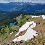 A scenic mountain landscape with patches of melting snow on a rocky slope in the foreground. Below, a small turquoise alpine lake is surrounded by dense evergreen forest. In the distance, tall rugged peaks rise above the tree line under a partly cloudy sky