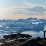 Landscape shot of Greenland ice sheets in the background and rocks in the foreground. Two polar explorers stand on top of the rocks gazing out into the polar sheets. 