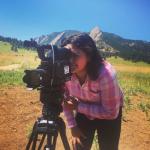 Person in a pink plaid shirt operating a video camera on a tripod in an open field with mountains in the background under a clear blue sky.