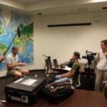 Group of students filming an interview indoors; one person holds a boom microphone, another operates a camera, while a man sits in front of them speaking, with a large world map on the wall behind