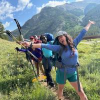 A group of happy hikers, dressed in outdoor gear and backpacks, poses enthusiastically on a scenic mountain trail. The front woman has her arms spread wide, smiling brightly, while the others behind her do the same, holding up trekking poles. They are surrounded by green grass, wildflowers, and majestic tree-covered mountains under a partly cloudy blue sky. The mood is joyful and energetic, capturing the spirit of adventure.