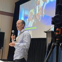 Ryan Vachon presenting at the Sustainable Research + Innovations Congress. There is a camera on the right side of the screen and a presentation on the back. Ryan is holding a microphone which he is speaking into