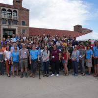 A large group of participants from the Resilient Futures Teacher Workshop, held in June 2025, pose together outdoors in front of a historic stone building with red-tiled roofs and arched windows. The diverse group includes men and women of various ages, many wearing name badges and casual attire. The setting appears to be a university campus, with a white event tent and clear skies in the background.  