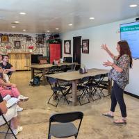 A woman is presenting to a small group of adults seated at tables in a bright, modern classroom. She is pointing at a large screen displaying a slide titled “Climate Action Bingo.” The audience appears engaged, with some taking notes or looking at handouts. The room includes posters, bookshelves, and various educational materials, creating a welcoming learning environment.