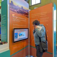 A person with long hair, wearing a gray jacket and black shorts, stands in front of an interactive exhibit display. The exhibit features an orange panel with white text and a mounted touchscreen showing informational content. The background includes another display and a window letting in daylight
