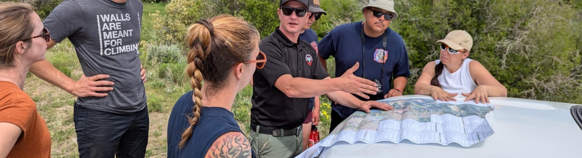 HEART Force Teacher Fellows and Earth Force Program Managers gather around the hood of a car in the field, studying a map with Theo Engel, Assistant Fire Management Officer from the Gunnison Ranger District. The group, surrounded by shrubs, learns about the 2023 Lowline Fire. 