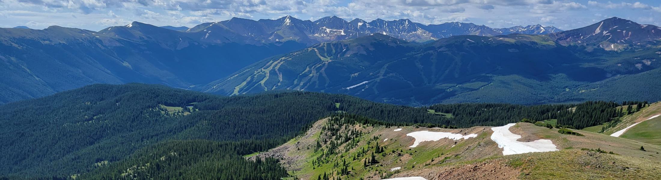Photo from on top of a mountain range with blue lake at the bottom and mountain range in the back during summer