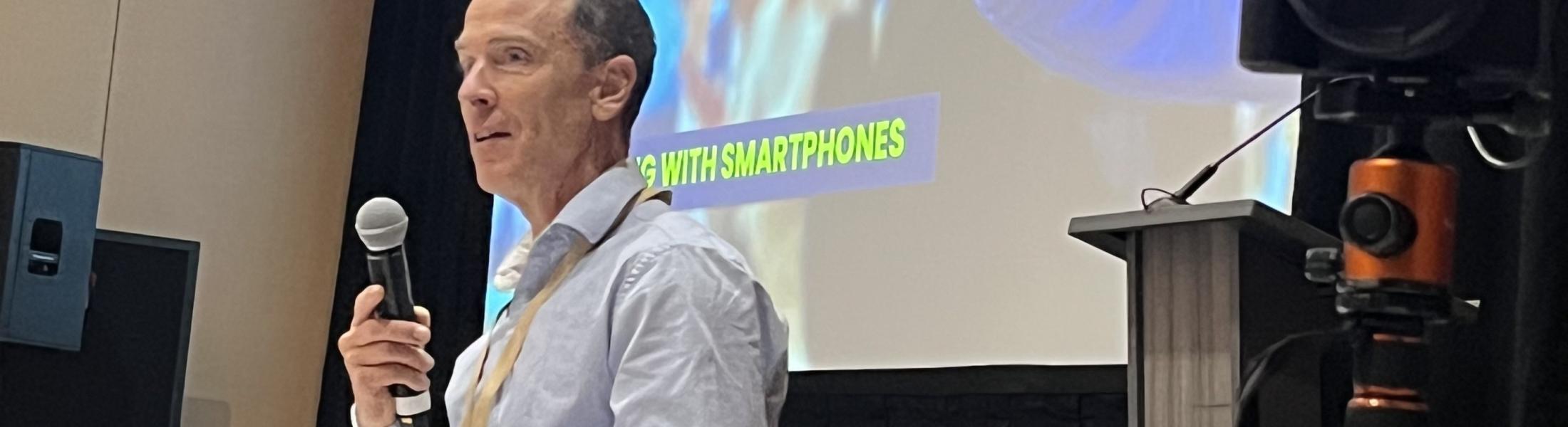 Ryan Vachon presenting at the Sustainable Research + Innovations Congress. There is a camera on the right side of the screen and a presentation on the back. Ryan is holding a microphone which he is speaking into