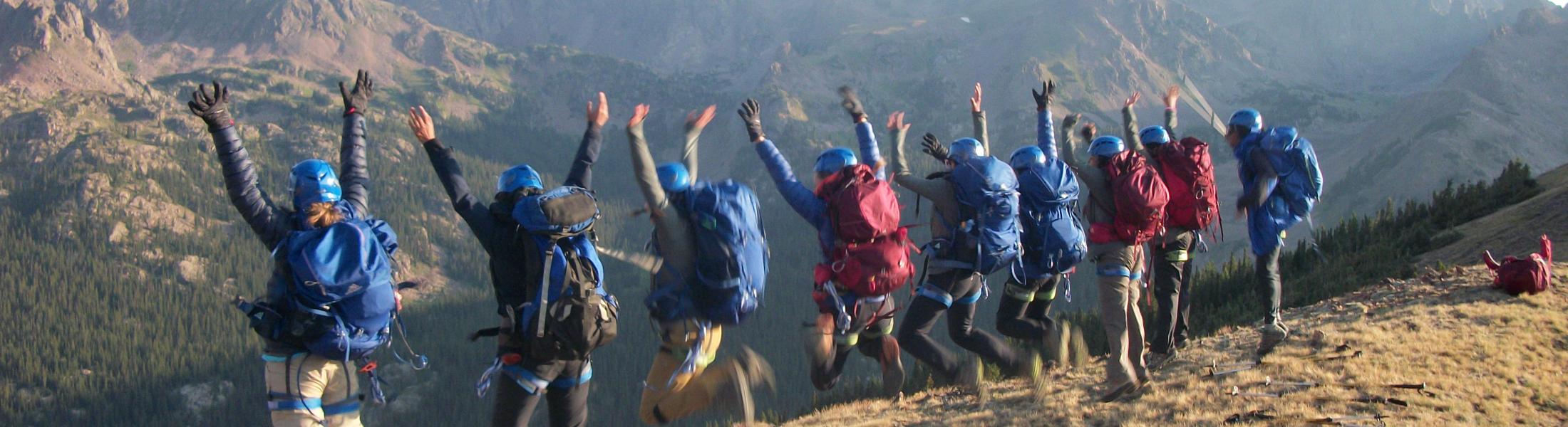 Girls* on Rock participants 2022 A group of hikers wearing helmets and large backpacks jump into the air with their arms raised in celebration on a mountain ridge. They are overlooking a vast range of rugged mountains with forested valleys below, under a clear sky. Trekking poles are scattered on the ground in the foreground.