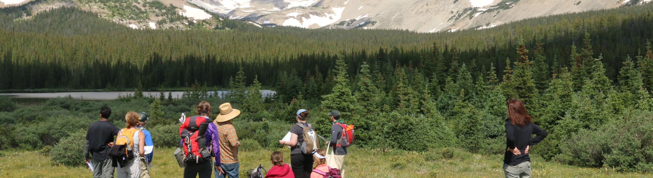 RECCS people A group of hikers stands and sits on grassy terrain, facing a scenic view of snow-dusted mountains and dense evergreen forest under a partly cloudy sky. Some people have backpacks, and one person wears a wide-brimmed hat. A small lake is visible at the base of the forest, adding to the tranquil alpine landscape.