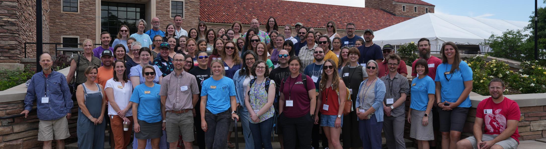 A large group of participants from the Resilient Futures Teacher Workshop, held in June 2025, pose together outdoors in front of a historic stone building with red-tiled roofs and arched windows. The diverse group includes men and women of various ages, many wearing name badges and casual attire. The setting appears to be a university campus, with a white event tent and clear skies in the background.  