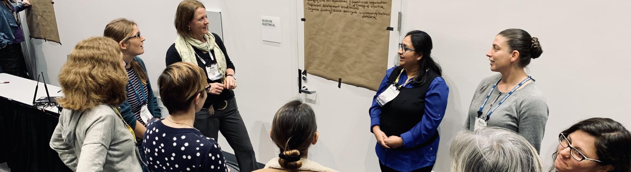 A group of women are gathered in a conference or workshop setting, standing in a circle around a large piece of brown paper taped to a white wall. The paper contains handwritten notes, and two women appear to be leading the discussion. The room has gray carpeting with red and blue lines, and there are more posters on the walls. The participants appear engaged and are wearing name tags and casual attire