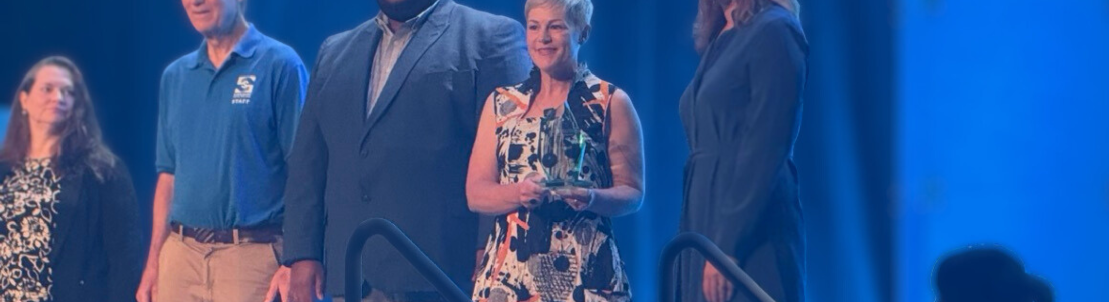 Carrie McDougall stands on stage with peers and award presenters at the ASTC Award Ceremony as she is presented with her trophy celebrating her achievement.