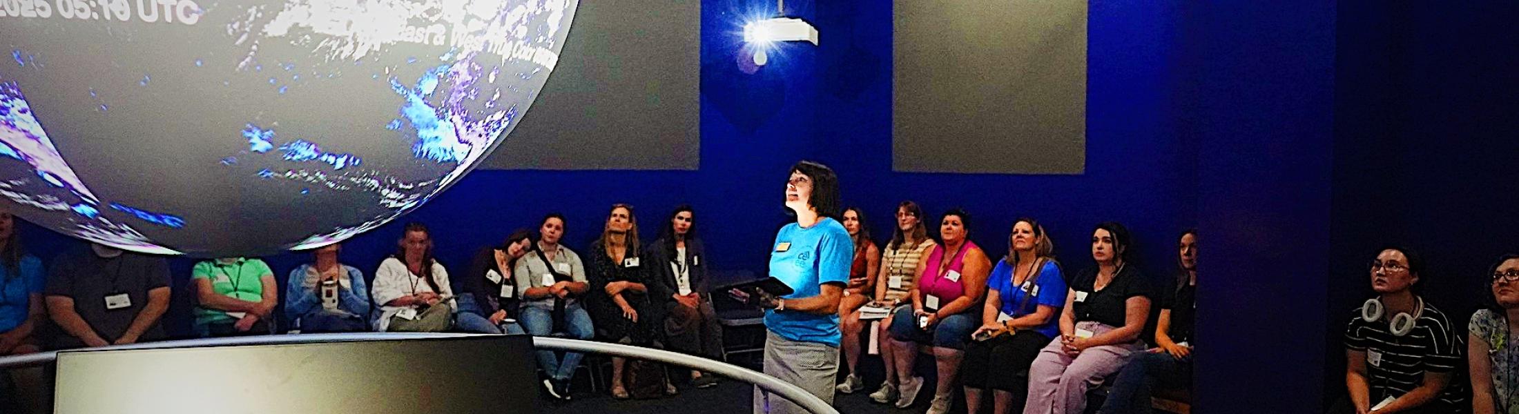A group of people sit in a dark, blue-walled room watching a presentation. A large illuminated globe display hangs from the ceiling, showing Earth with cloud patterns and text overlays. A presenter in a blue shirt stands near the globe, holding a tablet, while the audience observes attentively