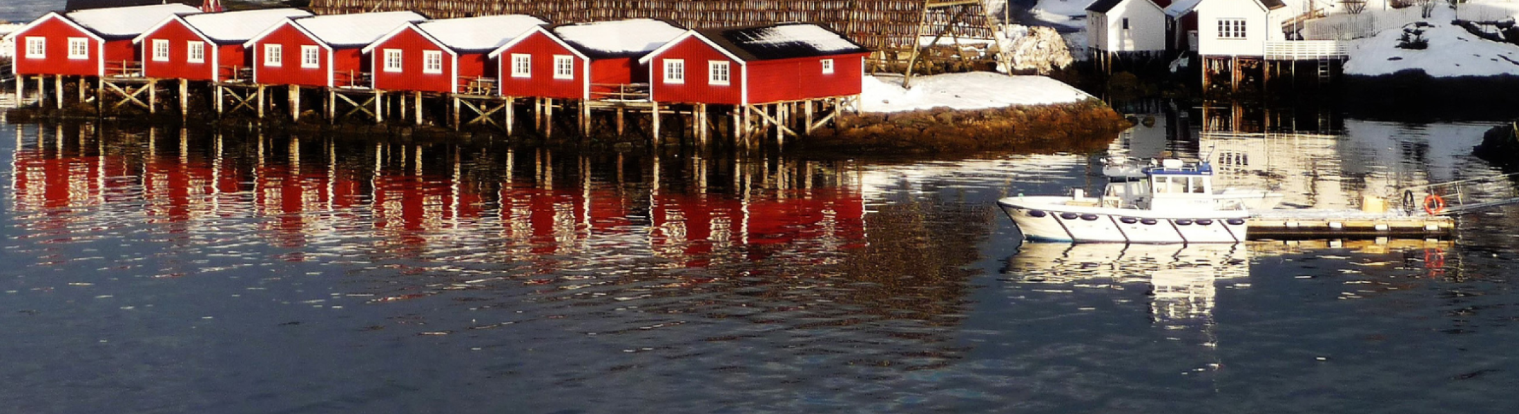 Row of traditional red fishermen’s cabins on stilts along a snowy shoreline in Norway, with a wooden fish-drying rack behind them, white houses on the right, and a small boat docked on calm reflective water
