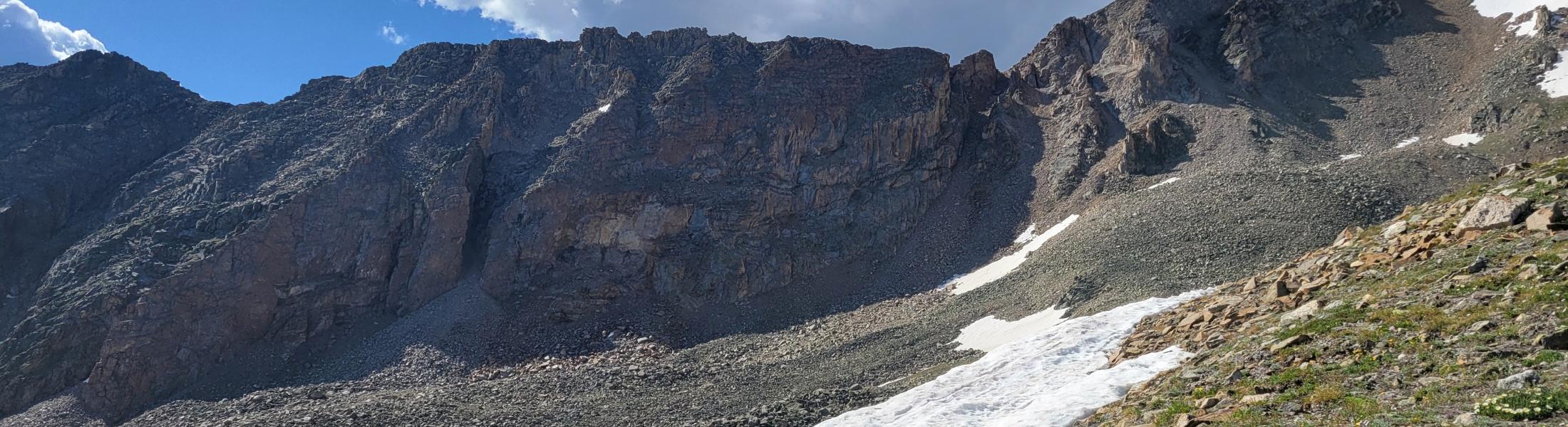 Mountain landscape with rocky peaks under a bright blue sky with scattered white clouds. A patch of snow stretches across the slope in the foreground, surrounded by green grass and scattered rocks