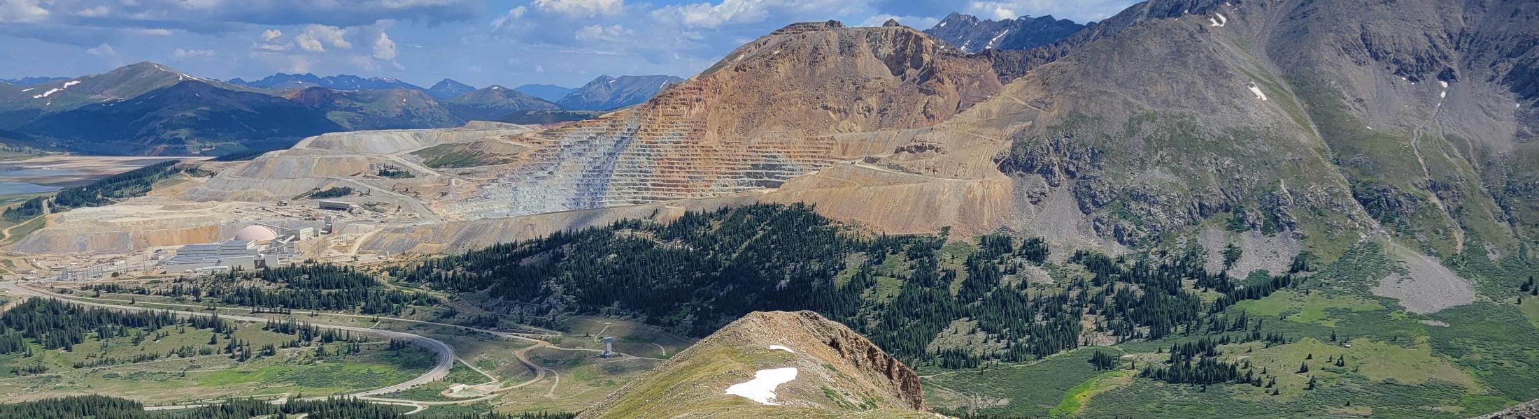 Expansive mountain landscape with rugged peaks in the background and a valley of green forests and meadows below. A narrow ridge runs through the center with patches of snow along its slope under a bright blue sky with scattered clouds