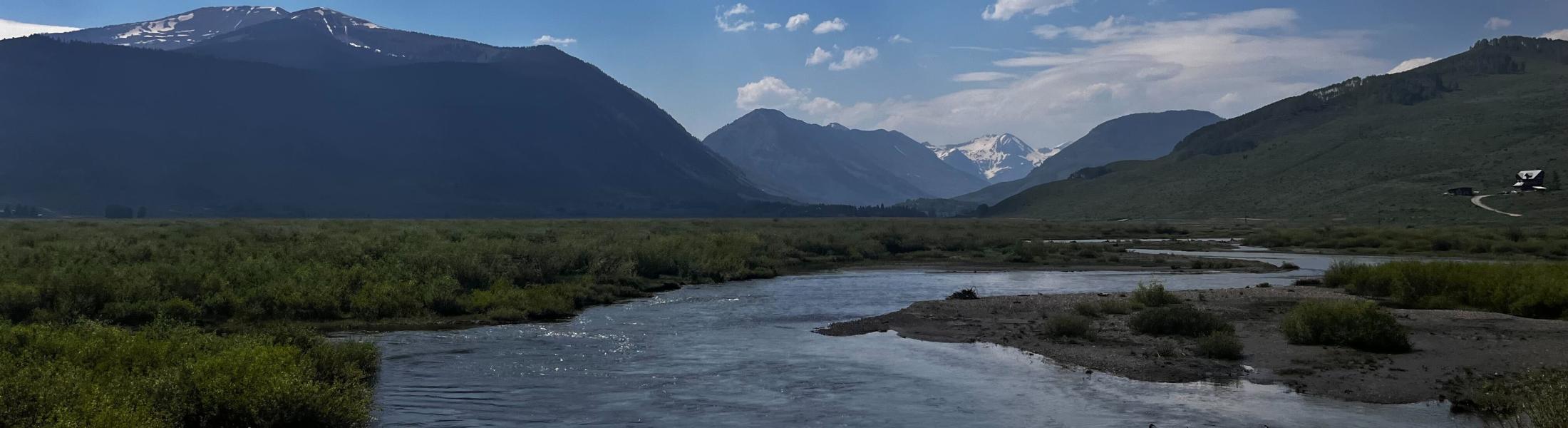 Wide river flowing through a green valley with dense shrubs and grasses in the foreground. Snow-capped mountains rise in the distance under a partly cloudy blue sky, with darker forested slopes and open hillsides on either side