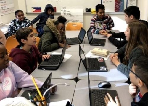 A group of students using laptops at desks all pushed together to form one collaborative workspace.