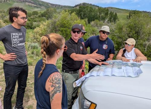 HEART Force Teacher Fellows and Earth Force Program Managers gather around the hood of a car in the field, studying a map with Theo Engel, Assistant Fire Management Officer from the Gunnison Ranger District. The group, surrounded by shrubs, learns about the 2023 Lowline Fire. 