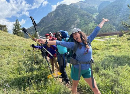 A group of happy hikers, dressed in outdoor gear and backpacks, poses enthusiastically on a scenic mountain trail. The front woman has her arms spread wide, smiling brightly, while the others behind her do the same, holding up trekking poles. They are surrounded by green grass, wildflowers, and majestic tree-covered mountains under a partly cloudy blue sky. The mood is joyful and energetic, capturing the spirit of adventure.