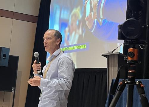 Ryan Vachon presenting at the Sustainable Research + Innovations Congress. There is a camera on the right side of the screen and a presentation on the back. Ryan is holding a microphone which he is speaking into