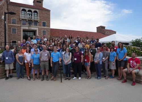 A large group of participants from the Resilient Futures Teacher Workshop, held in June 2025, pose together outdoors in front of a historic stone building with red-tiled roofs and arched windows. The diverse group includes men and women of various ages, many wearing name badges and casual attire. The setting appears to be a university campus, with a white event tent and clear skies in the background.  