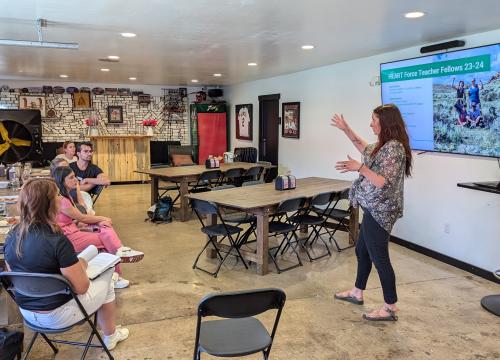 A woman is presenting to a small group of adults seated at tables in a bright, modern classroom. She is pointing at a large screen displaying a slide titled “Climate Action Bingo.” The audience appears engaged, with some taking notes or looking at handouts. The room includes posters, bookshelves, and various educational materials, creating a welcoming learning environment.