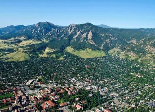 Aerial view of Boulder, Colorado, showing the city with its grid-like streets surrounded by lush greenery, with the Flatirons rock formations and mountains in the background under a clear blue sky