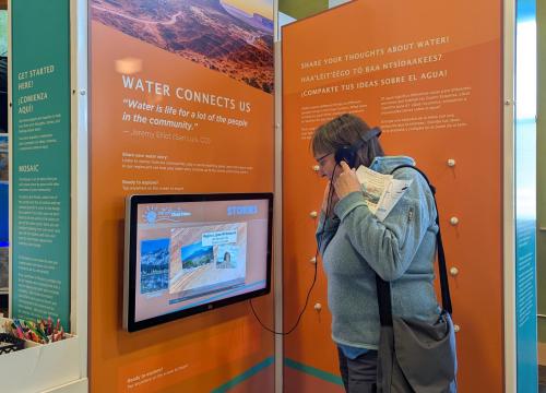 A person with long hair, wearing a gray jacket and black shorts, stands in front of an interactive exhibit display. The exhibit features an orange panel with white text and a mounted touchscreen showing informational content. The background includes another display and a window letting in daylight