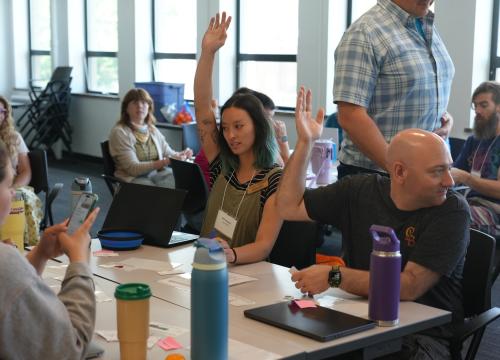 A group of adults sit around tables in a bright classroom setting. One woman, seated in the center, raises her hand while others listen and look on. Water bottles, notebooks, and laptops are spread across the tables, suggesting a workshop or professional learning session