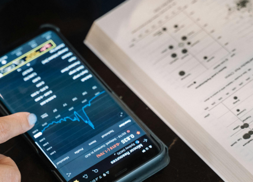 A hand scrolls on a phone aside a textbook.