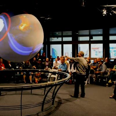 Person presenting an SOS exhibit with the SOS globe hanging from the ceiling and a rail around it. Lots of people in the back learning about SOS