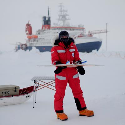 Person standing on an arctic ice field in a red suit with a boat behind them 