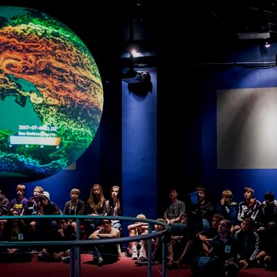 Beth Russel giving an SOS presentation to a large crowd of children. The SOS globe is hanging from the ceiling showing sea surface temperatures-green, blue, orange and yellow are shown on the globe