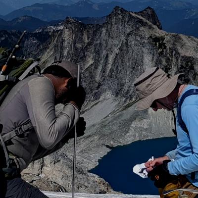 Two people conducting fieldwork on a mountain side with a blue lake below