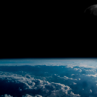  A view from space showing the Earth's curved horizon covered in a layer of clouds, with the dark expanse of space above. The Moon is partially visible in the upper right corner against the black sky.